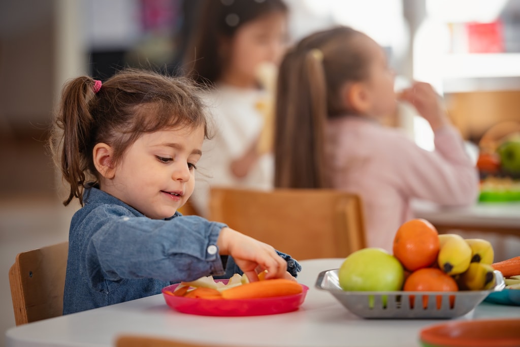 Young girl enjoys a healthy snack at preschool while other children sit nearby, promoting healthy eating habits, nutrition, and diversity in an early childhood classroom setting