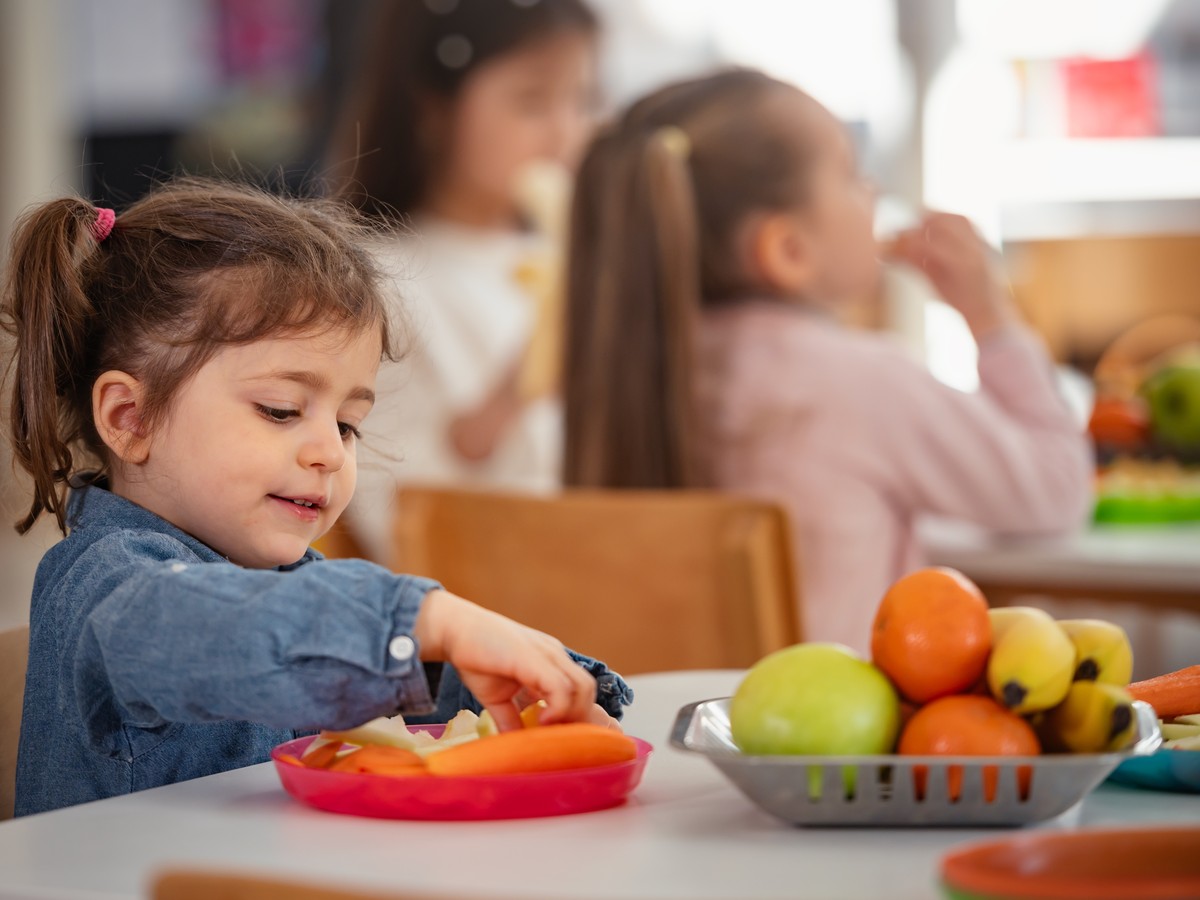 Young girl enjoys a healthy snack at preschool while other children sit nearby, promoting healthy eating habits, nutrition, and diversity in an early childhood classroom setting