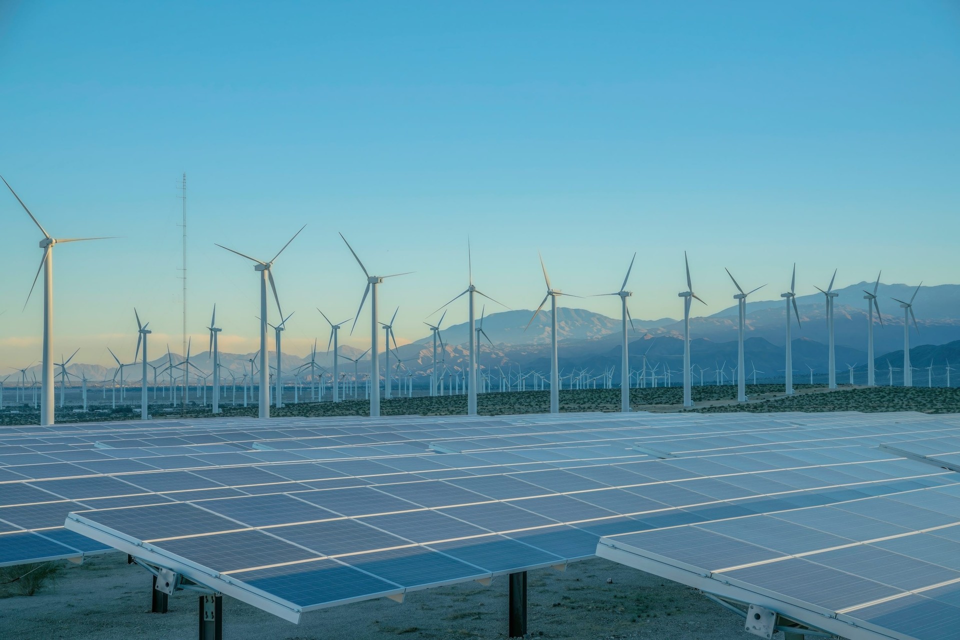 Solar panels and wind turbines against the sunset sky on a desert in California. There are solar panels and a view of a windmill farm at the back near the mountain range.