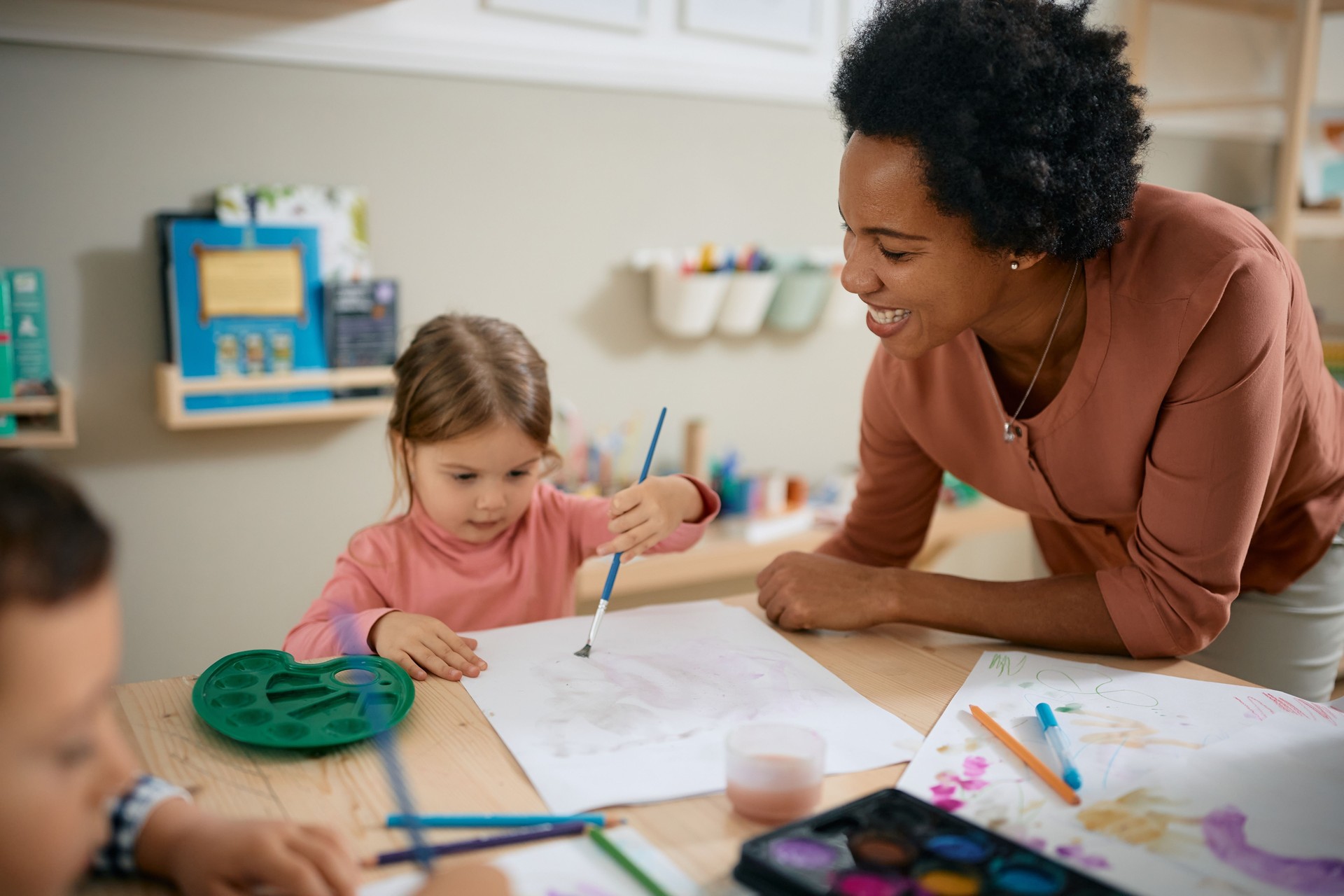 Happy African American art teacher assisting kids in coloring on the paper at preschool.