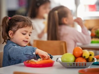 Young girl enjoys a healthy snack at preschool while other children sit nearby, promoting healthy eating habits, nutrition, and diversity in an early childhood classroom setting