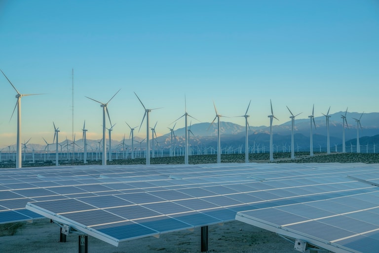 Solar panels and wind turbines against the sunset sky on a desert in California. There are solar panels and a view of a windmill farm at the back near the mountain range.