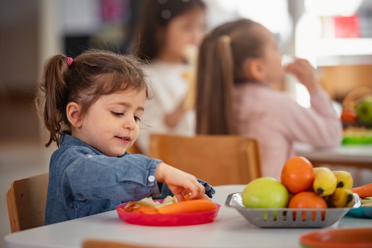 Young girl enjoys a healthy snack at preschool while other children sit nearby, promoting healthy eating habits, nutrition, and diversity in an early childhood classroom setting